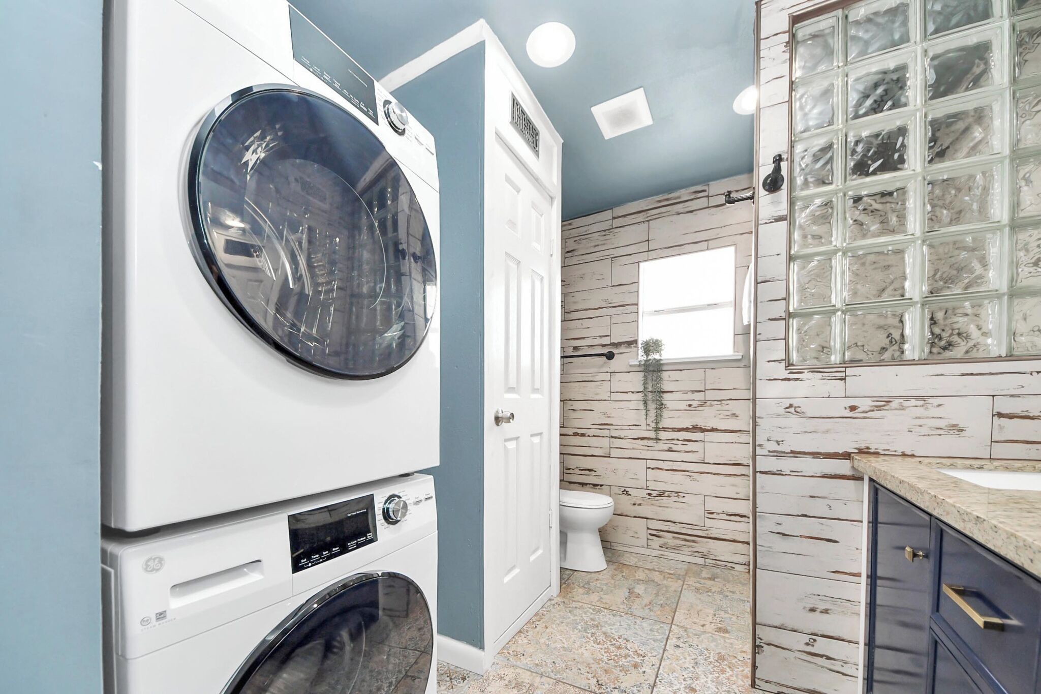 A laundry room with stacked washers and dryers, a white door, and a bathroom with a wooden wall and a glass shower enclosure.