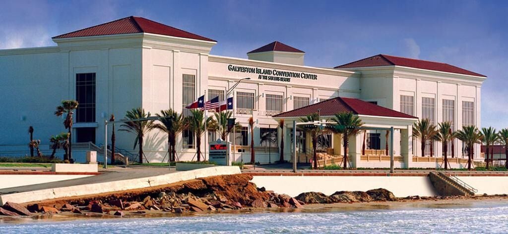 A large, white building with a red roof, named Galveston Island Convention Center, located near a body of water.