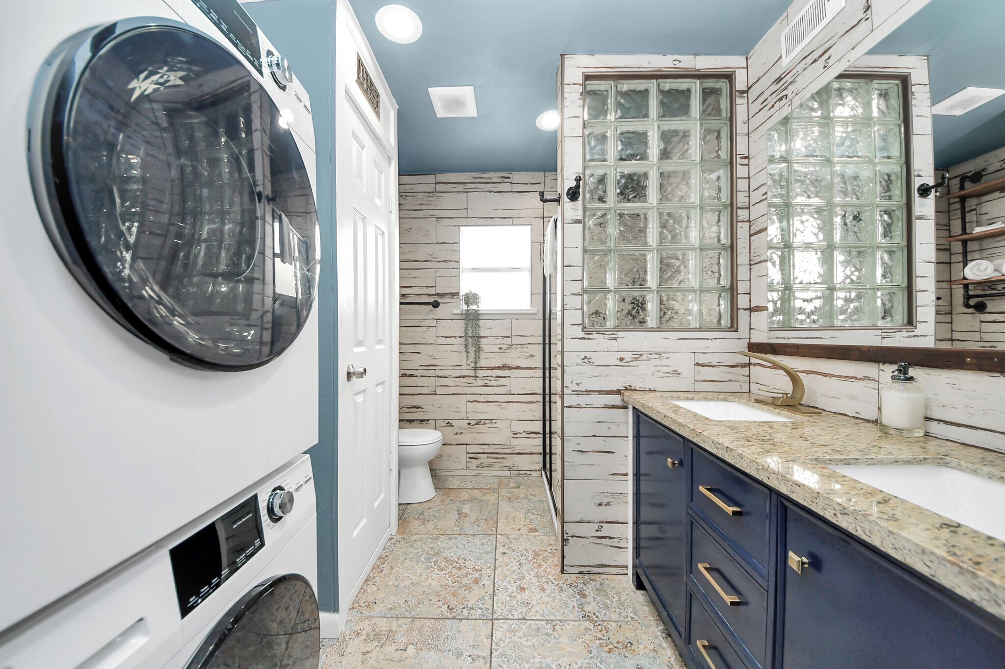 A compact laundry room with a front-loading washer and dryer, a white toilet, and a large window with frosted glass panels.