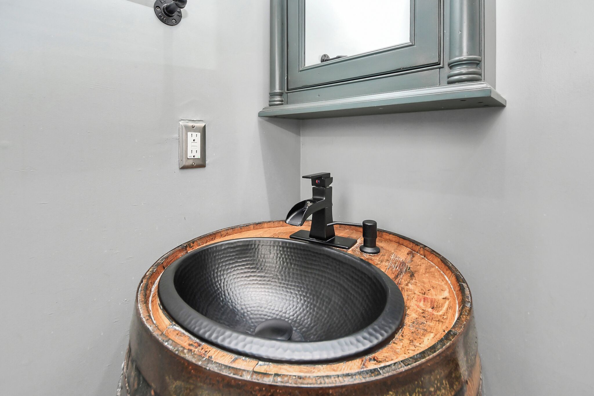 A bathroom with a round wooden sink, a black faucet, a mirror above the sink, and a light switch on the wall.