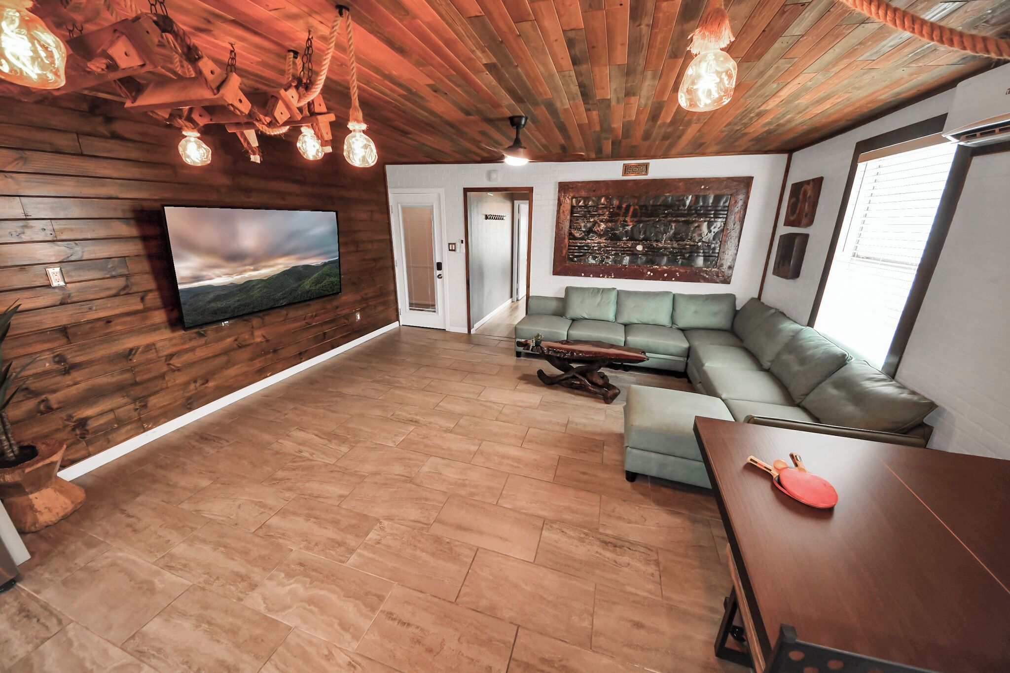A spacious living area with a wooden ceiling, a large flat-screen TV, a green sofa, and a dining table with a paddle.