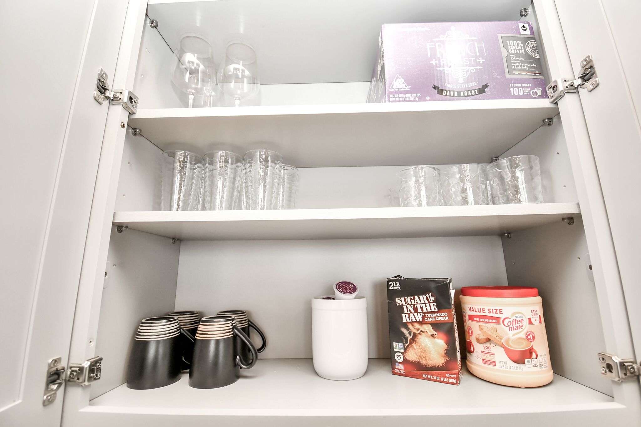 Ein Schrank mit Regalen, der klare Gläser, schwarze Tassen, eine weiße Tasse, eine Zuckerbox und ein Kaffeiglas enthält.
