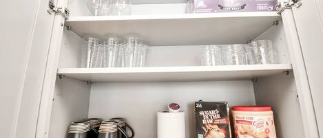 Ein Schrank mit Regalen, der klare Gläser, schwarze Tassen, eine weiße Tasse, eine Zuckerbox und ein Kaffeiglas enthält.