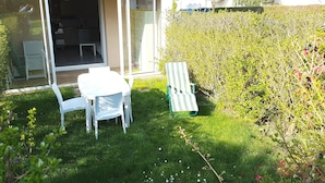 En uteplats med ett bord och stolar, en grön loungestol och en glasdörr som leder in till ett rum med en säng och en stol.