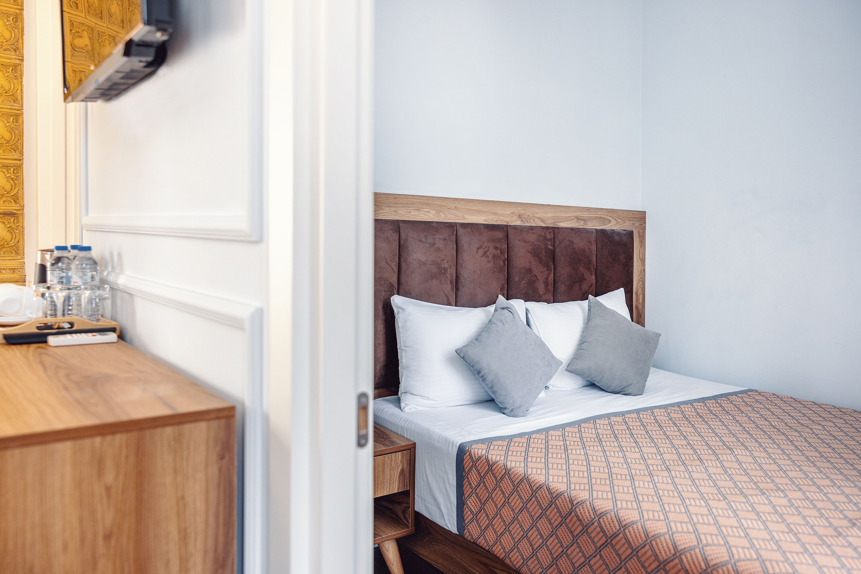 A neatly made bed with a wooden headboard, white bedding, and two gray pillows. A wooden nightstand with a lamp and a few bottles is beside the bed.