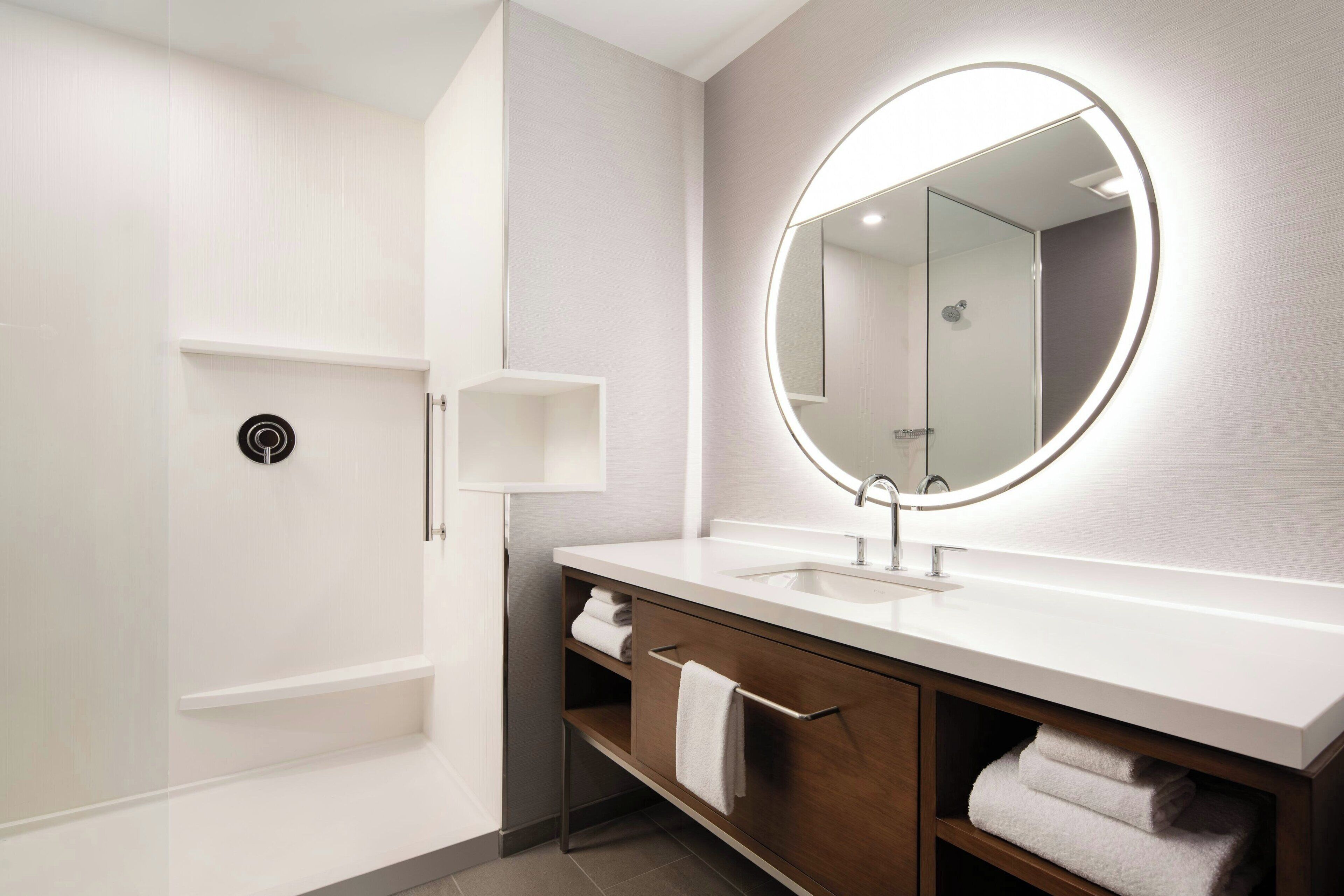 A modern bathroom with a white vanity, a large mirror with a circular light, and a shower area.