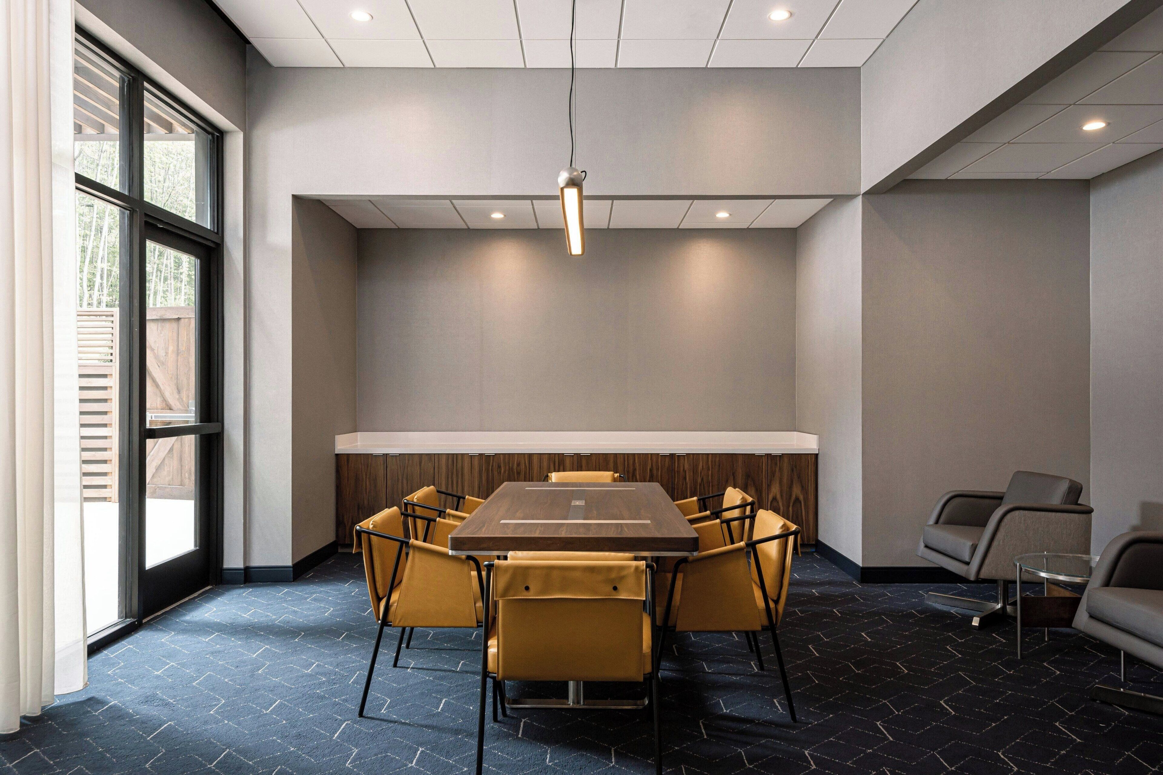 A conference room with a rectangular table, yellow chairs, a standing lamp, and a view through large windows.