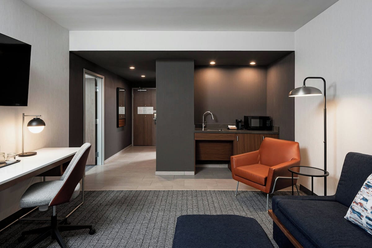 A modern hotel room with a desk, a sofa, a chair, a small kitchen area, and a TV mounted on the wall.