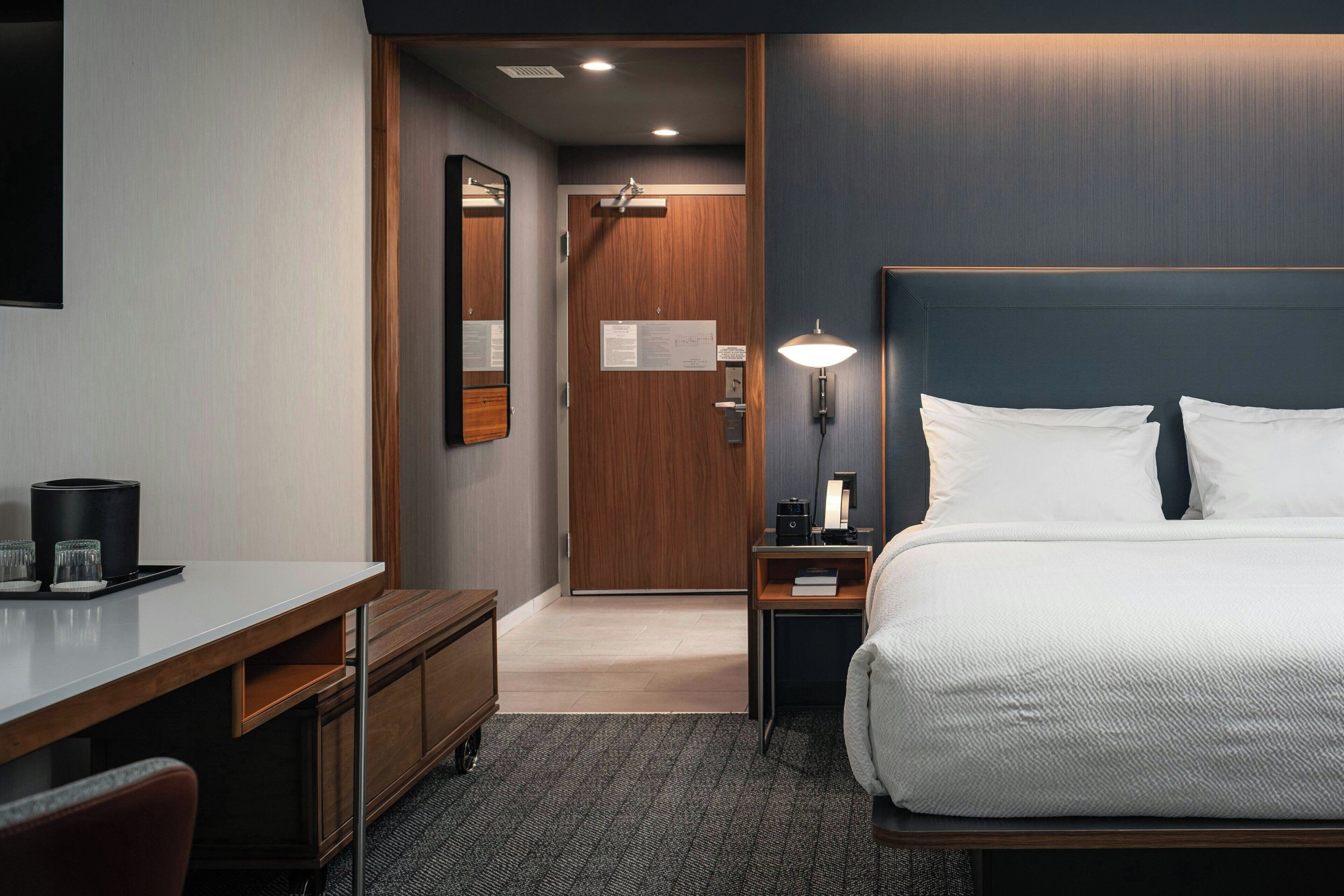Ein Hotelzimmer mit Bett, Schreibtisch, Stuhl, Nachttisch, Lampe und einer Tür zu einem anderen Raum.