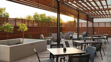 An outdoor patio area with tables and chairs, a wooden pergola, and a planter with small trees.