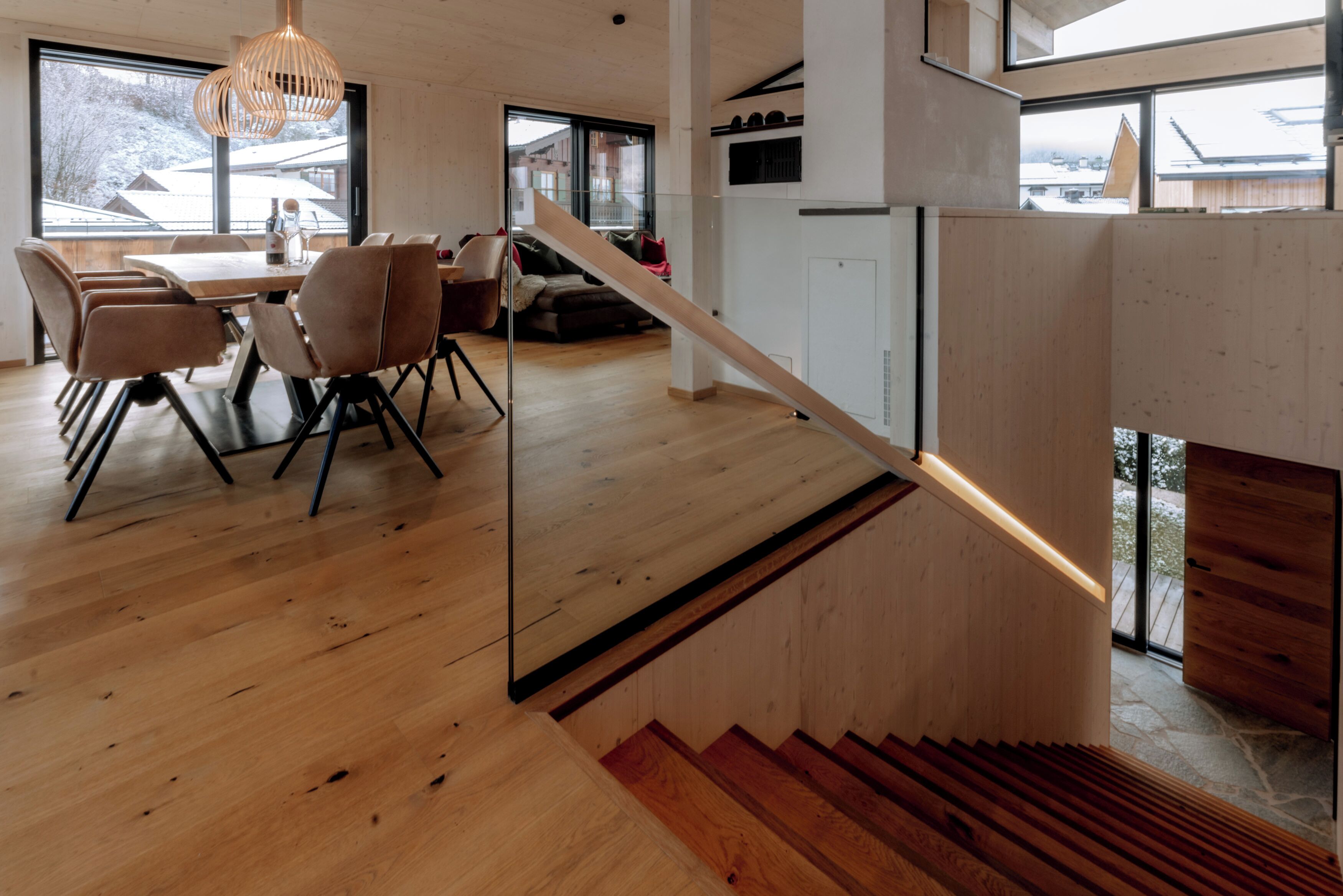 Et moderne interiør med et træspisebord og stole, en trappe med gelænder i træ, samt store vinduer der giver udsigt ud