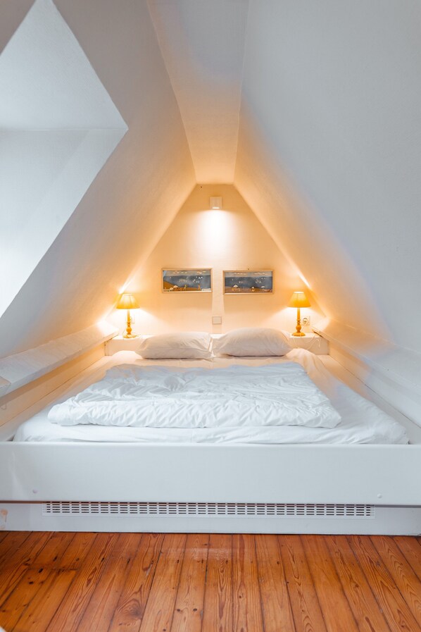 Un dormitorio con techo inclinado, una cama con ropa de cama blanca, dos lámparas de noche y un cuadro en la pared.