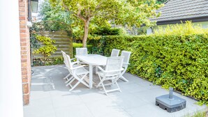 Un patio con mesa y sillas blancas, rodeado de follaje y un muro de ladrillo.