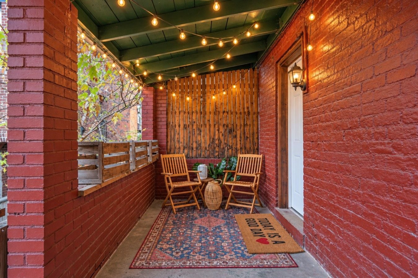 Un espace extérieur abrité, avec des murs en briques, des chaises en bois, un tapis à motifs et des guirlandes lumineuses.