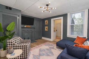 Een kamer met een open haard, een geruit bankje, een blauwe bank en een groot raam.