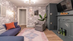 Un salon avec un canapé bleu, un tapis à motifs, un fauteuil à carreaux et une cheminée sur laquelle est posé un panneau indiquant « Papa ».