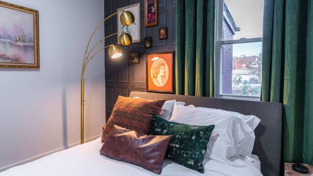 Une chambre à coucher avec un lit, des oreillers, une lampe sur pied et une vue sur la ville par la fenêtre.