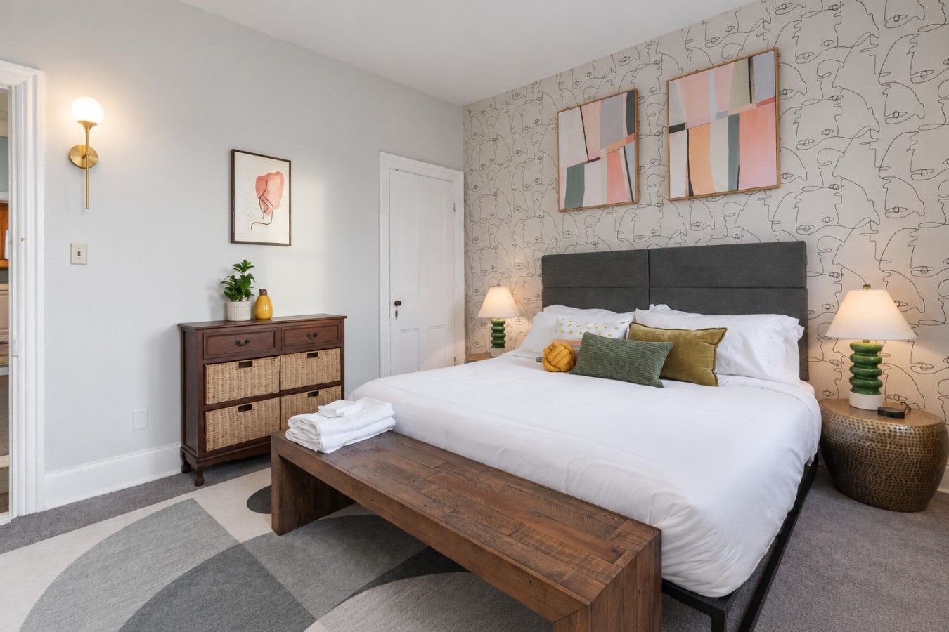 Ein Schlafzimmer mit einem Bett, Nachttischlampen, einem Nachttisch, einer Kommode, einer Bank und einer Wand mit abstrakter Kunst.