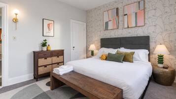 Une chambre à coucher comprenant un lit, des lampes de chevet, une table de chevet, une commode, un banc et un mur orné d’œuvres d’art abstraites.