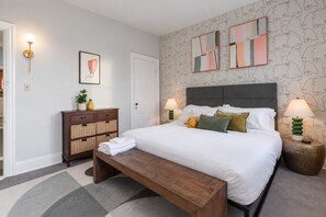Een slaapkamer met een bed, bedlampen, een nachtkastje, een dressoir, een bankje en een muur met abstracte kunst.