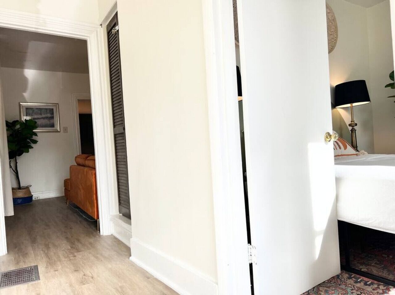 A hallway with a white door, a bed with a black headboard, a lamp, and a plant.