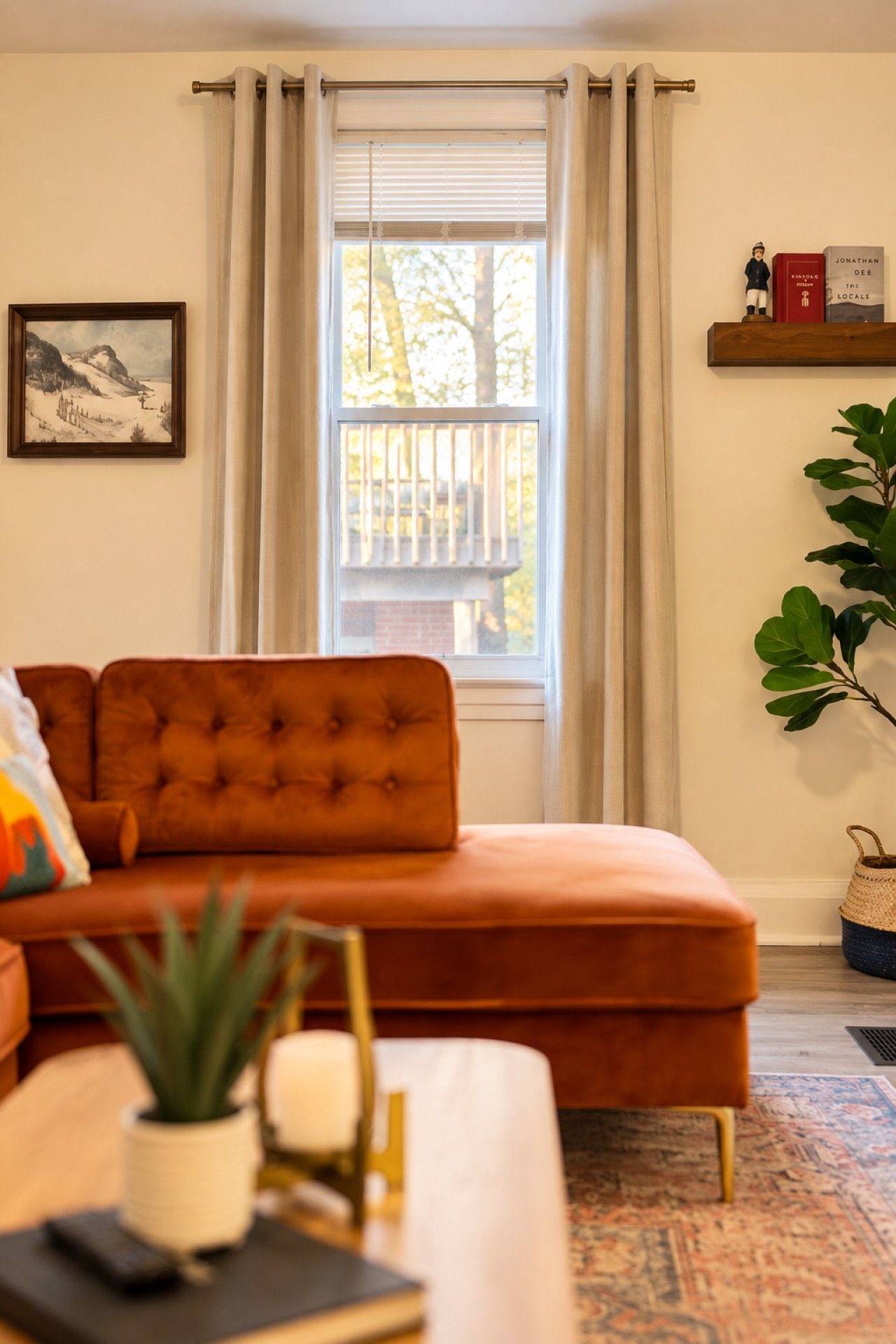 Ein Wohnzimmer mit einer Couch, einem Fenster mit Vorhängen, einem Gemälde und einem kleinen Bücherregal.