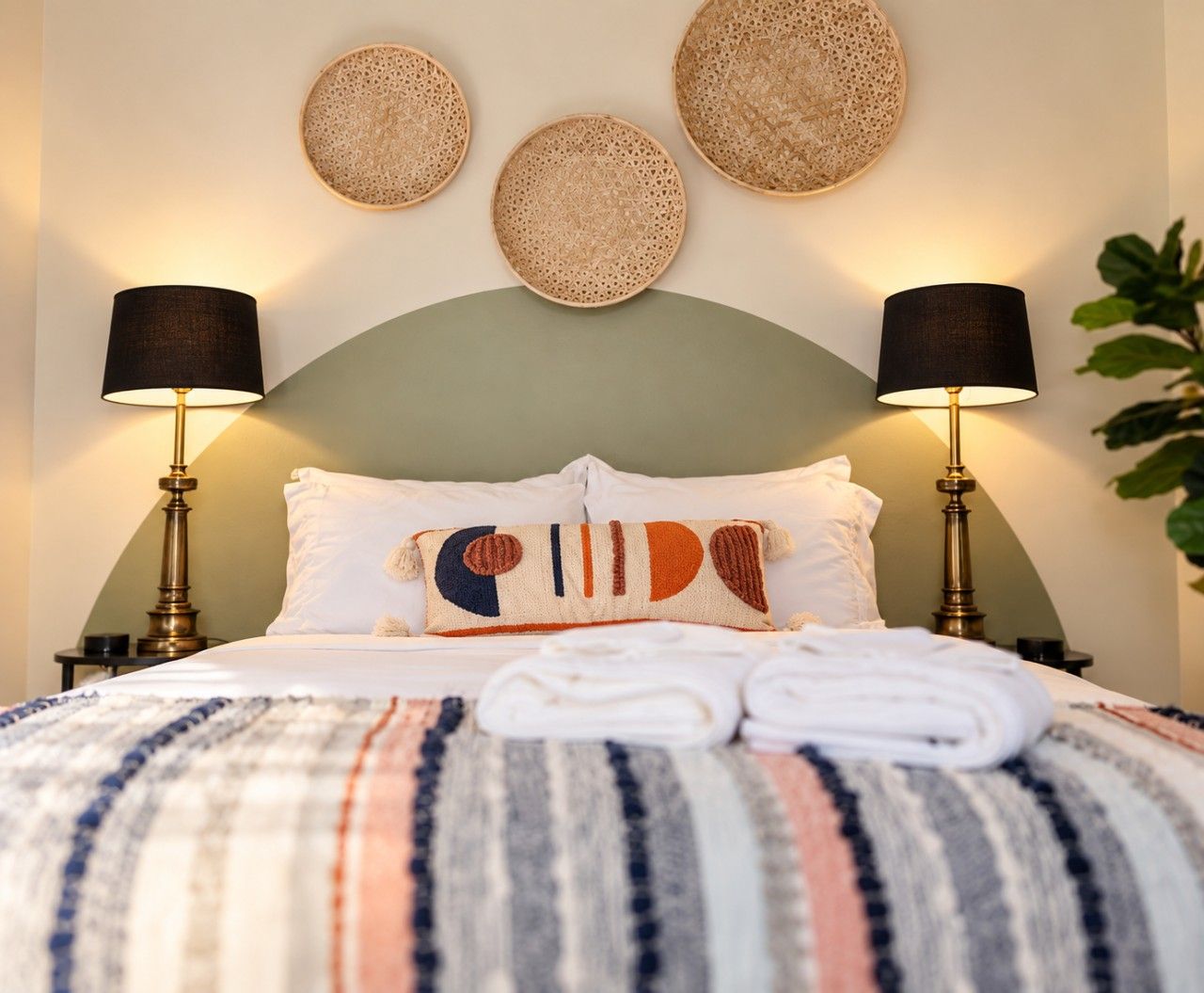 Cama bem arrumada com manta listrada, duas luminárias de apoio e uma parede decorada com três peças de arte em tecido.