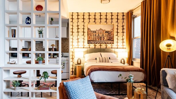 A hotel room with a large bed, a white shelving unit, a patterned wall, and a view of a building through the window.