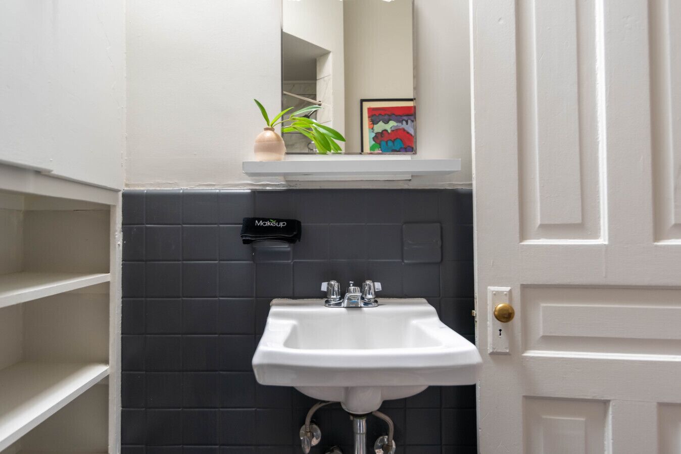 Un baño con un lavabo blanco, paredes de baldosas oscuras, un estante pequeño con una planta y un cuadro enmarcado, y un neceser colgado en la pared.