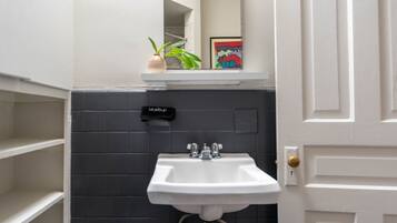 Un baño con un lavabo blanco, paredes de baldosas oscuras, un estante pequeño con una planta y un cuadro enmarcado, y un neceser colgado en la pared.