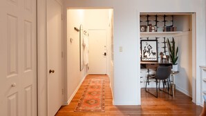 A hallway with a wooden floor, a patterned rug, a white door, and a desk with a chair and decorative items.