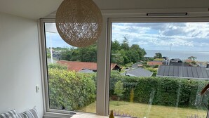 Ein Essbereich mit Holztisch und weißen Stühlen, großen Fenstern mit Blick auf einen Garten und Häuser sowie einer gewebten Leuchte.