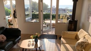 Ein Wohnzimmer mit Holzboden, einem runden Couchtisch mit Blumen, einer schwarzen Ledersofa, einem beigen Sofa und einem großen Fenster mit Blick ins Freie.