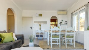 A living room with a sofa, a coffee table, and a dining area with a table and chairs. There is a door leading to another room and a window with curtains. - EDEN MAR 1A Roses (Rosas / Roses)