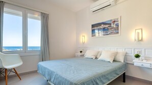 A bedroom with a bed, a chair, a nightstand, a wall-mounted air conditioner, a painting, and a view of the sea. - EDEN MAR 1A Roses (Rosas / Roses)
