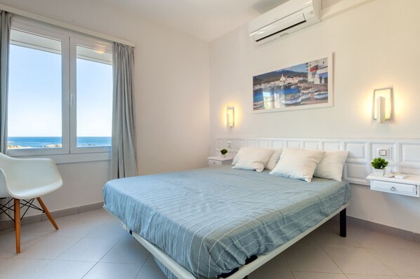 A bedroom with a bed, a chair, a nightstand, a wall-mounted air conditioner, a painting, and a view of the sea. - EDEN MAR 1A Roses (Rosas / Roses)
