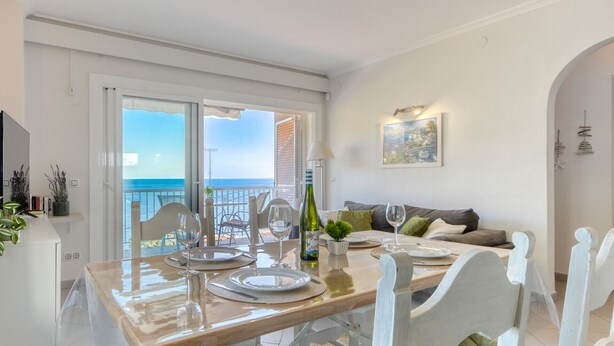 A dining area with a table set for four, a balcony with a view of the sea, and a living area with a sofa and a painting. - EDEN MAR 1A Roses (Rosas / Roses)