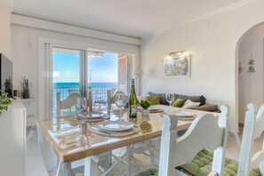 A dining area with a table set for four, a balcony with a view of the sea, and a living area with a sofa and a painting. - EDEN MAR 1A Roses (Rosas / Roses)