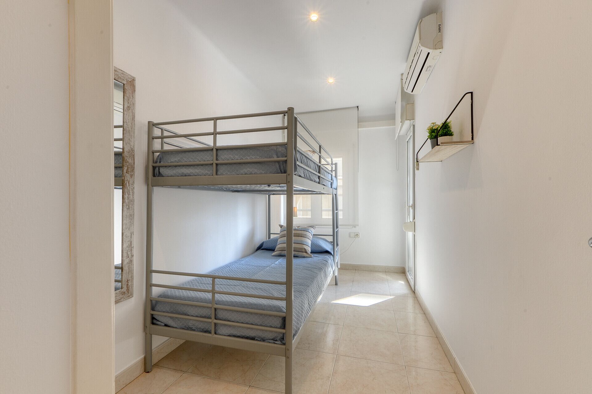 Dormitorio con literas, ventana, aire acondicionado de pared y una pequeña estantería con una planta.