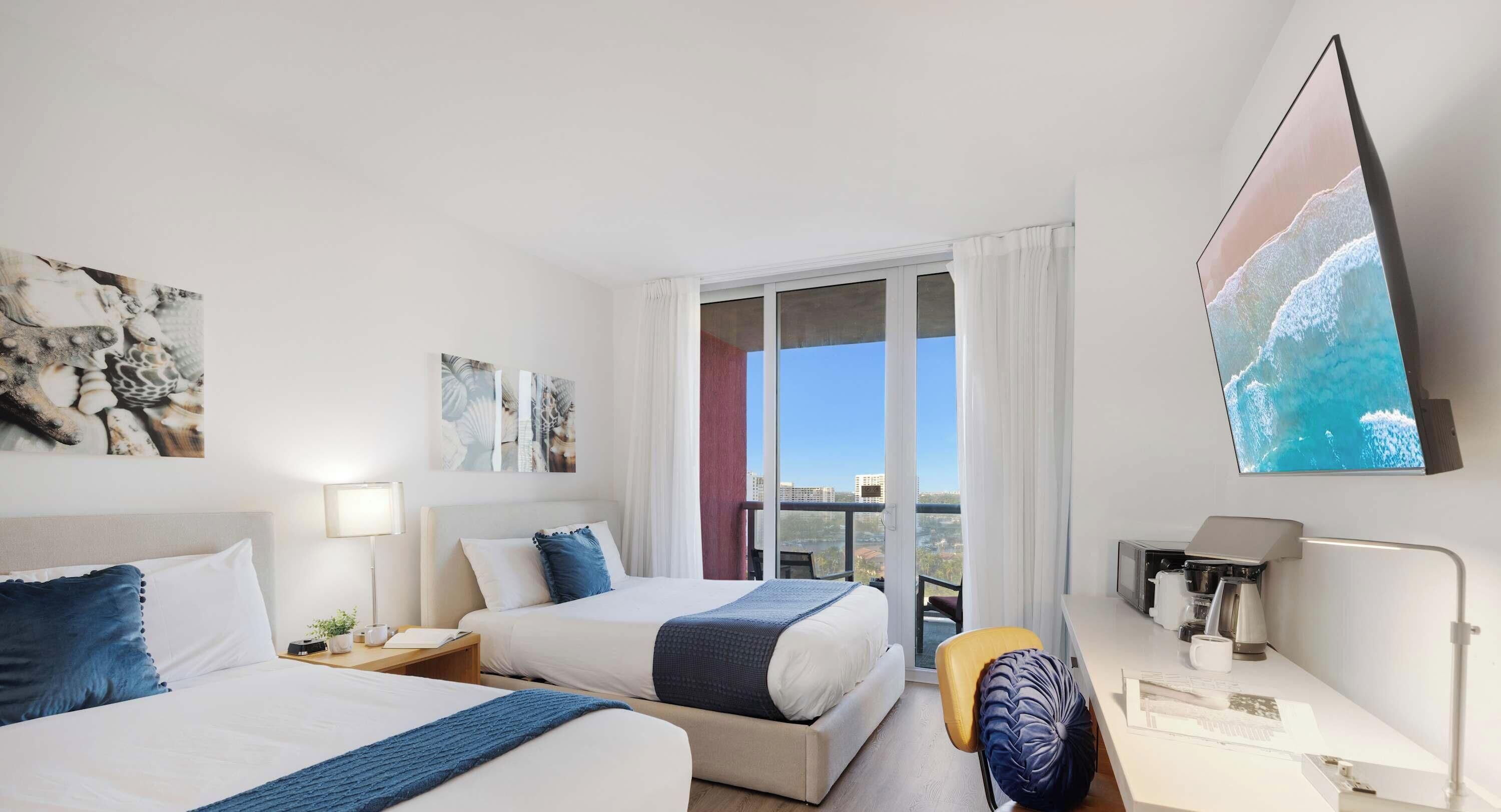 A hotel room with two beds, a desk, a chair, and a balcony with a city view.