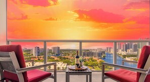 A balcony with two red chairs, a table with a bottle and glasses, and a cityscape view.