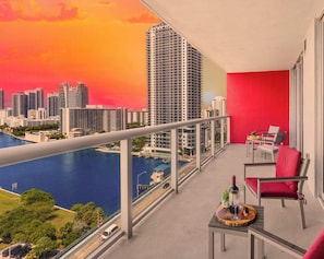 A balcony with a city view, a table with a bottle and glass, and red chairs.