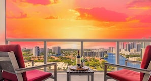 A balcony with two red chairs, a table with a bottle and glasses, and a cityscape view.