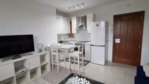Una cocina compacta con muebles blancos, horno empotrado y refrigerador. Un televisor de pantalla plana está montado en un mueble blanco, y una pequeña mesa de comedor con sillas se ubica frente a la cocina.