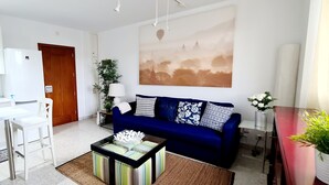 Una sala moderna con sofá azul, mesa de comedor blanca y área de cocina con armarios blancos y una campana de acero inoxidable.