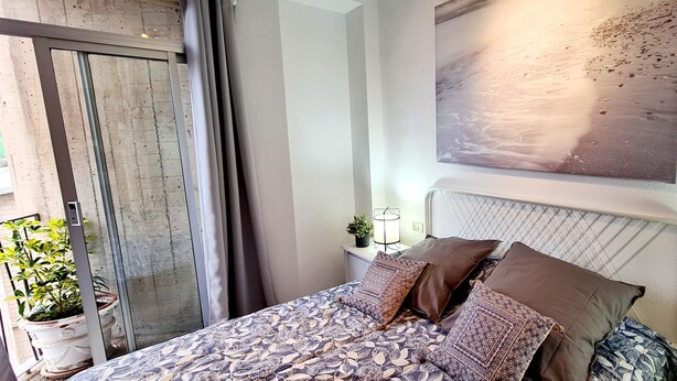 Un dormitorio con cama, almohadas, una mesita de noche, una planta en maceta y una obra de arte colgada en la pared.