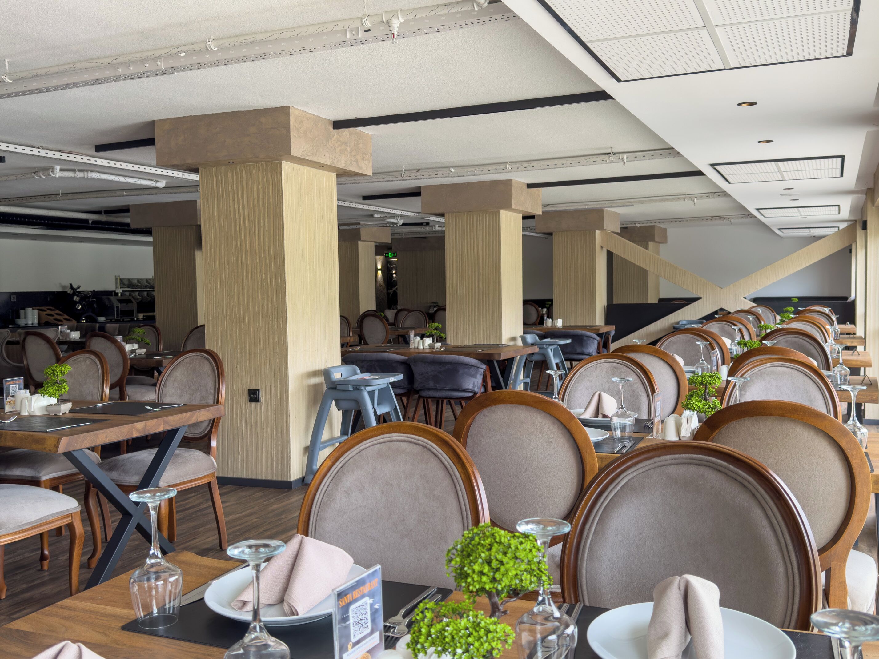 Un restaurante moderno con techos altos, conductos expuestos y una mezcla de sillas de madera y tapizadas. Las mesas están dispuestas con platos, vasos y pequeñas macetas.