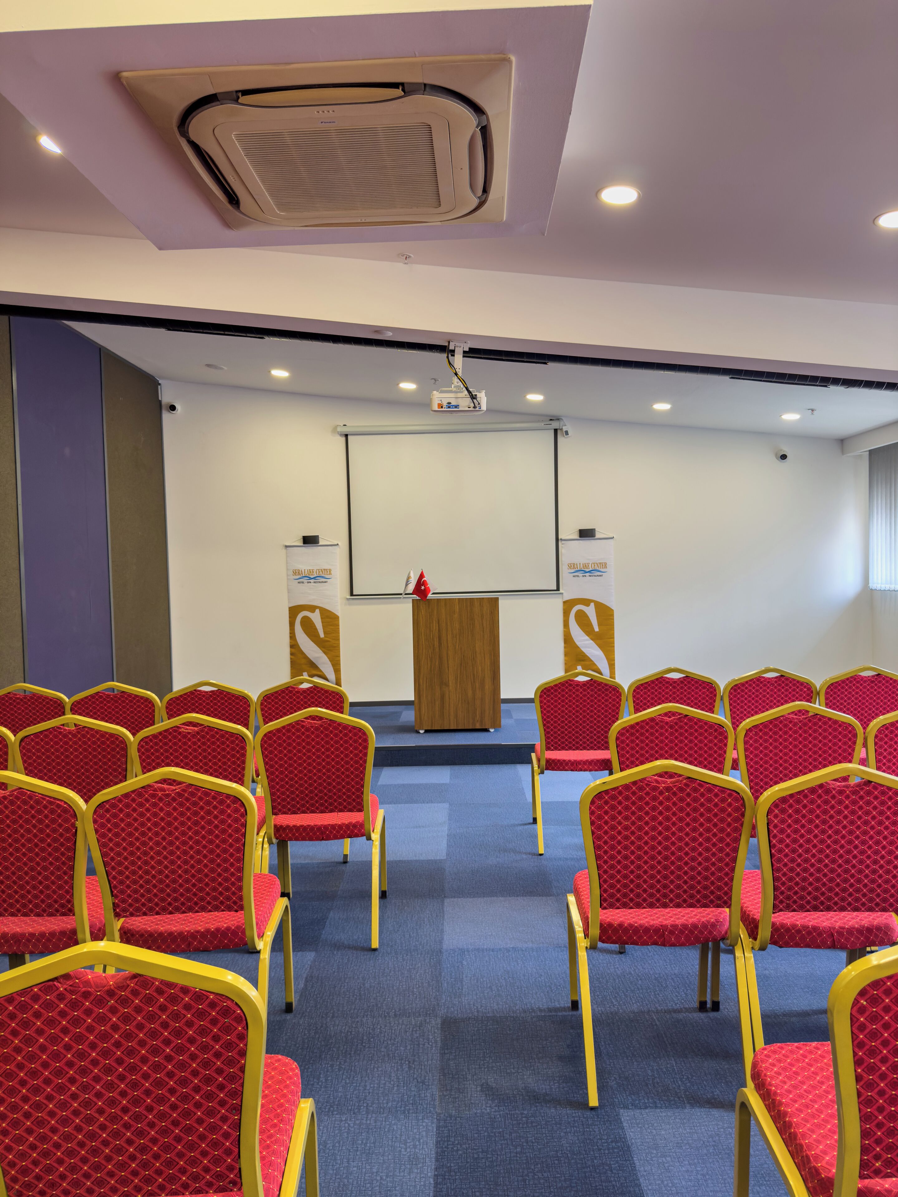 A conference room with rows of red chairs, a podium, and a projector screen.
