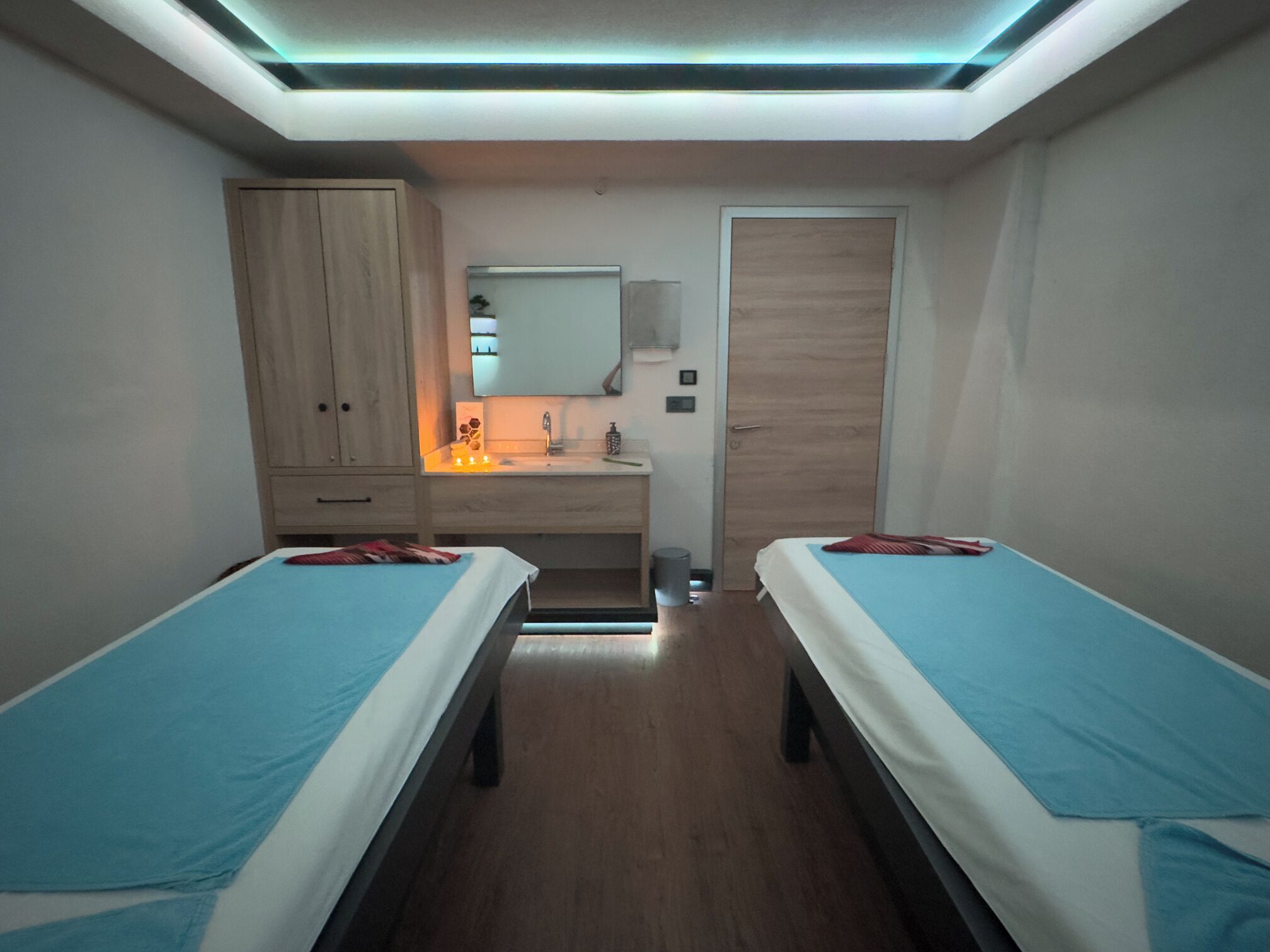 A room with two single beds, a wooden wardrobe, a mirror, a sink, and a door.