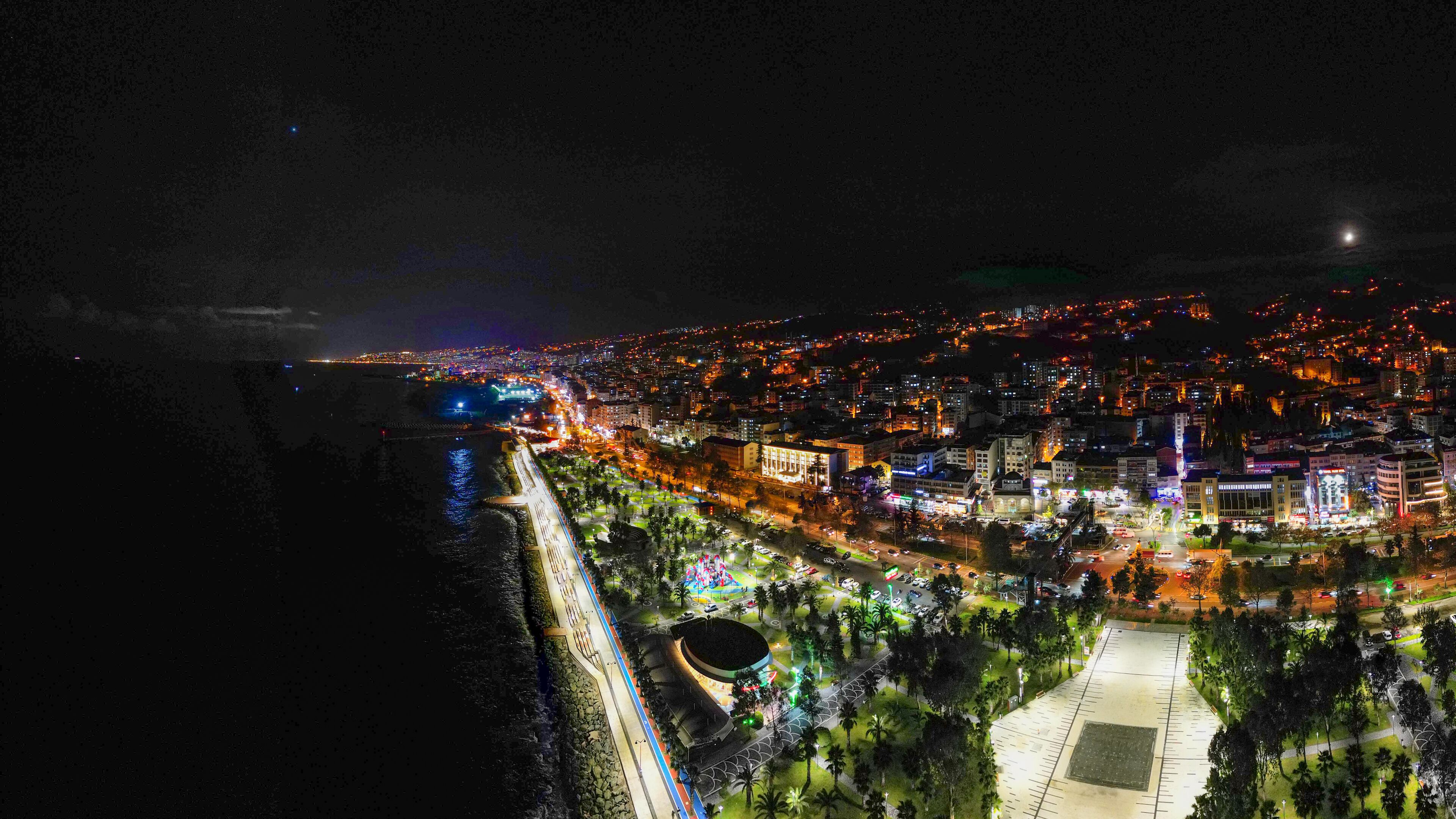 An aerial view of a city at night with illuminated buildings, a waterfront, and a large open plaza with palm trees.